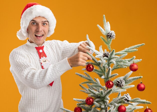 Impressed young handsome guy wearing christmas hat and santa claus tie standing near christmas tree decorating it with christmas ball ornament looking at camera isolated on orange background