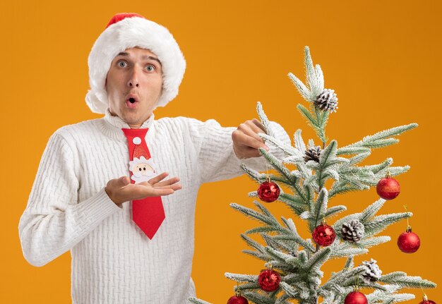 impressed young handsome guy wearing christmas hat and santa claus tie standing near christmas tree decorating it with christmas ball ornament looking  showing empty hand isolated on orange wall