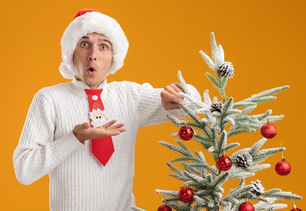 クリスマスの帽子とサンタ クロースのネクタイを着て感銘を受けた若いハンサムな男がクリスマス ツリーの近くに立って、オレンジ色の壁に孤立した空の手を見せてクリスマス ボールの飾りで飾っています。