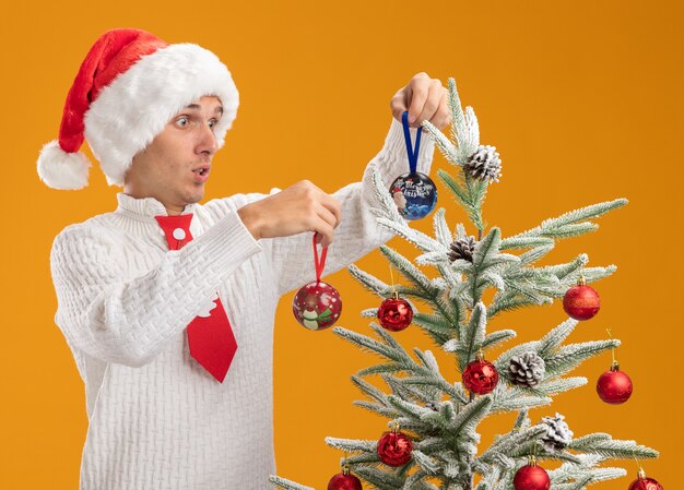 오렌지 배경에 고립 된 나무를보고 크리스마스 공 장식품으로 장식 크리스마스 트리 근처에 서 크리스마스 모자와 산타 클로스 넥타이를 입고 감동 젊은 잘 생긴 남자