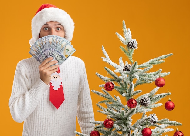 Free photo impressed young handsome guy wearing christmas hat and santa claus tie standing near decorated christmas tree holding money  from behind it isolated on orange wall