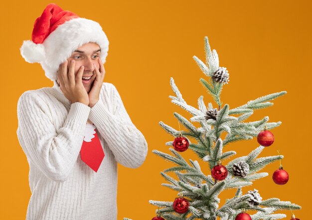 Impressed young handsome guy wearing christmas hat and santa claus tie standing near decorated christmas tree keeping hands on face looking down isolated on orange wall