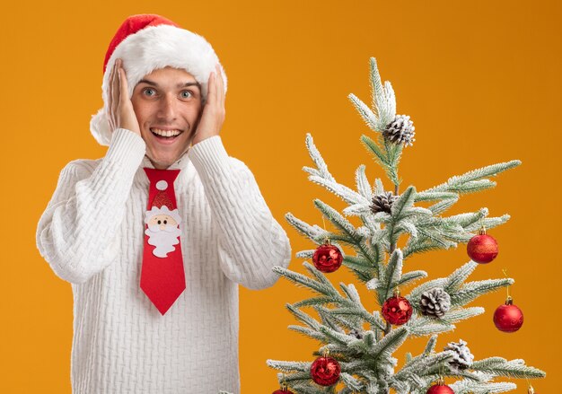 オレンジ色の背景で隔離のカメラを見て頭に手を置いて飾られたクリスマスツリーの近くに立っているクリスマス帽子とサンタクロースのネクタイを身に着けている感動の若いハンサムな男