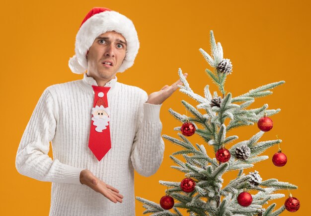 Впечатленный молодой красивый парень в рождественской шапке и галстуке санта-клауса, стоящий возле украшенной елки, указывая на нее руками, изолированными на оранжевой стене