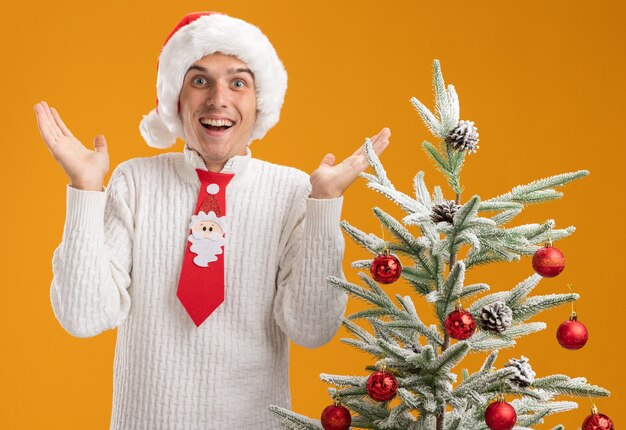 Impressed young handsome guy wearing christmas hat and santa claus tie standing near decorated christmas tree  showing empty hands isolated on orange wall