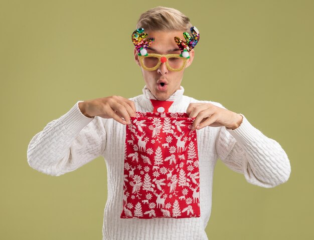 impressed young handsome guy wearing christmas novelty glasses and santa claus tie holding opening christmas sack looking inside it isolated on olive green wall