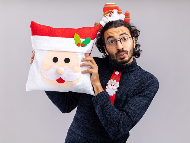 Impressed young handsome guy wearing christmas tie with hair hoop holding christmas pillow around face isolated on white background