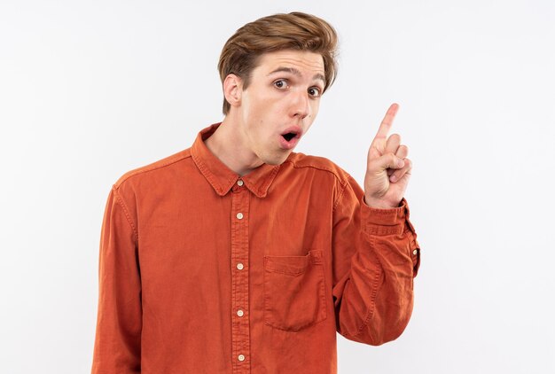 Impressed young handsome guy wearing red shirt points at up isolated on white wall