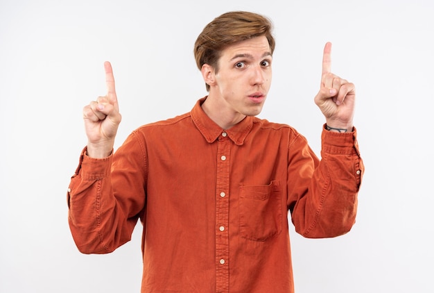 Impressed young handsome guy wearing red shirt points at up isolated on white wall