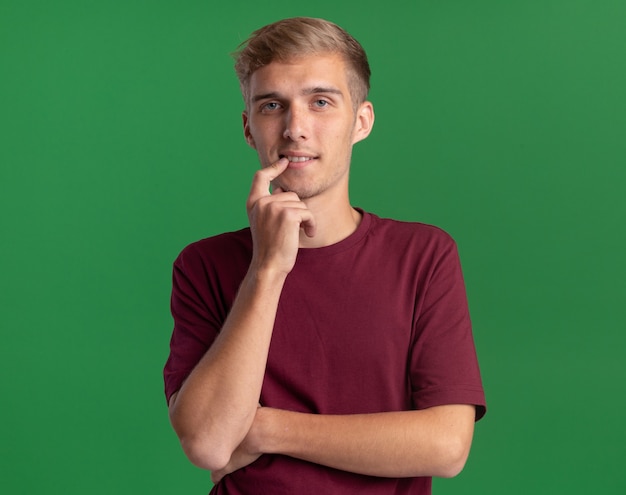 Foto gratuita giovane ragazzo bello colpito che indossa la camicia rossa che mette il dito sulla bocca isolata sulla parete verde