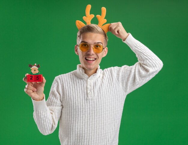 緑の壁に分離された日付をつかむヘッドバンドとクリスマスツリーのおもちゃを保持しているメガネとトナカイの角のヘッドバンドを身に着けている感動の若いハンサムな男
