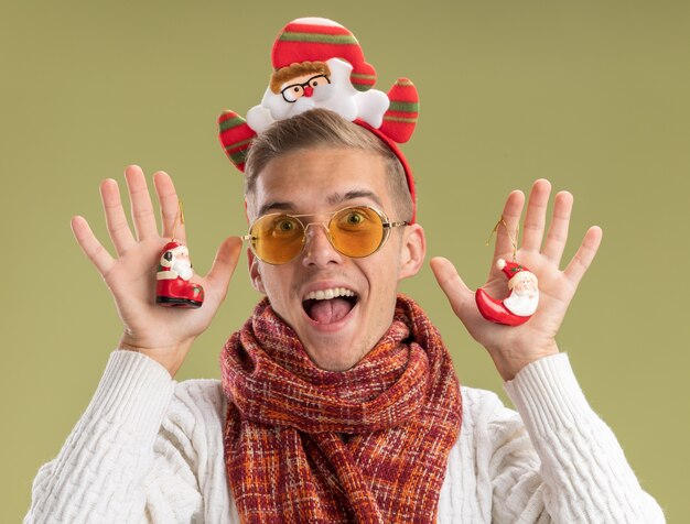 Impressed young handsome guy wearing santa claus headband and scarf looking at camera holding santa claus christmas ornaments looking at camera isolated on olive green background