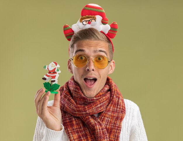 Impressed young handsome guy wearing santa claus headband and scarf looking at camera holding santa claus toy isolated on olive green background