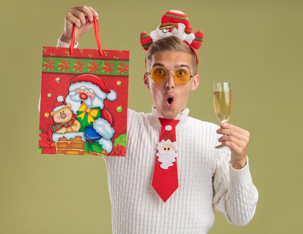 Впечатленный молодой красивый парень в повязке на голову санта-клауса и галстуке держит бокал шампанского и поднимает рождественский подарочный пакет, изолированный на оливково-зеленой стене