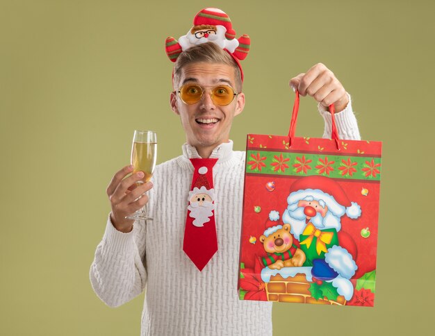 Impressed young handsome guy wearing santa claus headband and tie looking at camera holding glass of champagne and christmas gift bag isolated on olive green background