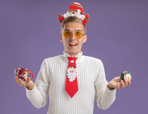 サンタクロースのヘッドバンドを身に着けている印象的な若いハンサムな男と紫色の壁に分離されたクリスマスボールの飾りを保持している眼鏡