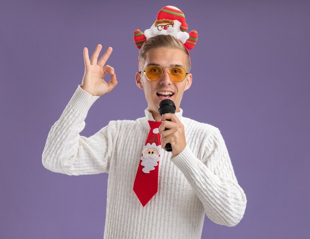 サンタクロースのヘッドバンドを身に着けて、紫色の壁に分離されたokサインをしている口の近くでマイクを保持している眼鏡でネクタイをしている印象的な若いハンサムな男