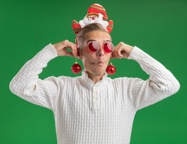 Impressed young handsome guy wearing santa claus headband with glasses hanging christmas baubles on ears looking at side with pursed lips isolated on green background