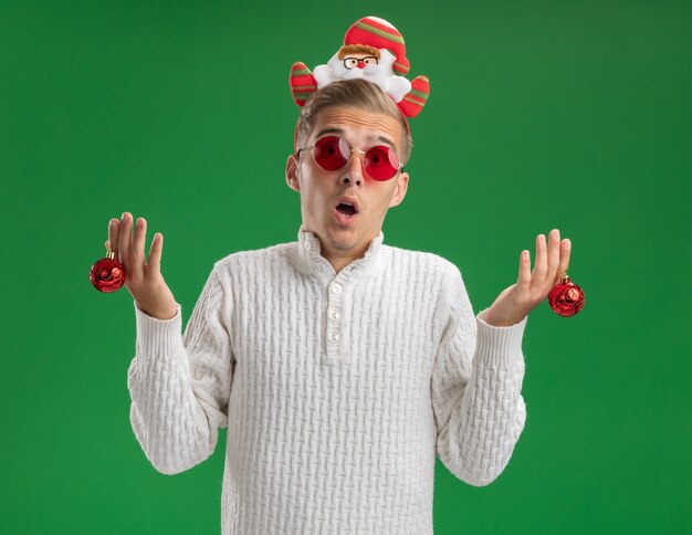 Impressed young handsome guy wearing santa claus headband with glasses holding christmas ornament balls  isolated on green wall