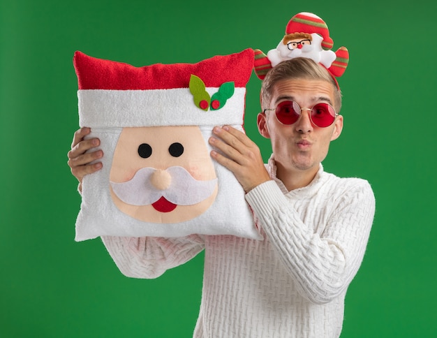 Free photo impressed young handsome guy wearing santa claus headband with glasses holding santa claus pillow looking at camera doing kiss gesture isolated on green background