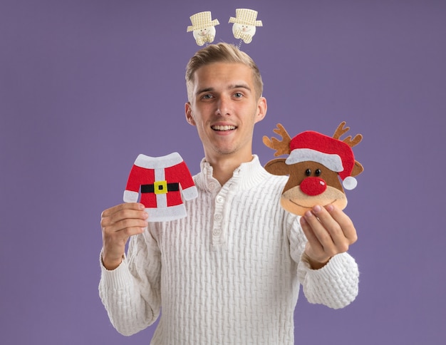 Free photo impressed young handsome guy wearing snowman headband holding and stretching out christmas paper ornaments towards  isolated on purple wall