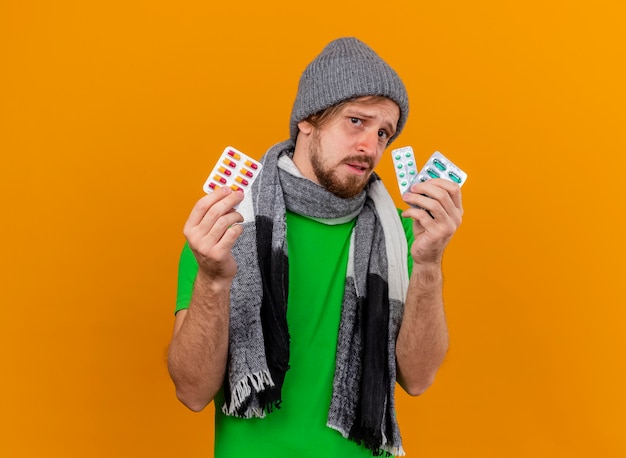 Impressed young handsome ill man wearing winter hat and scarf holding packs of capsules looking at front isolated on orange wall