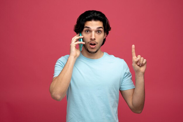 впечатленный молодой красивый мужчина смотрит в камеру, указывая вверх, разговаривая по телефону на красном фоне