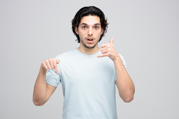 Impressed young handsome man looking at camera showing call gesture pointing down isolated on white background