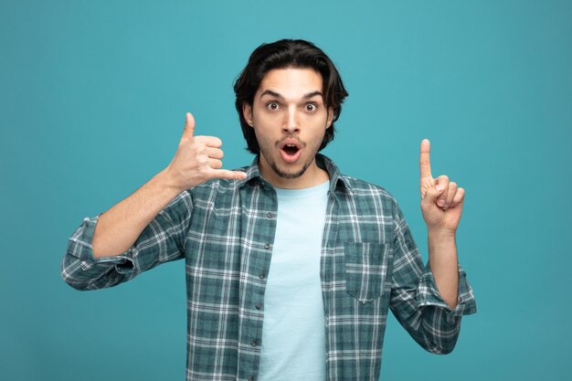 impressed young handsome man looking at camera showing call gesture pointing up isolated on blue background