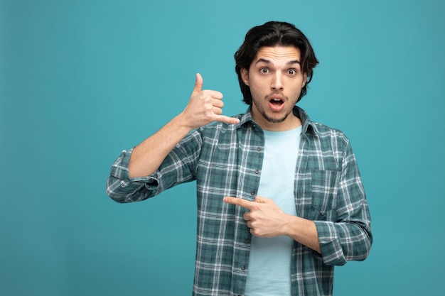 impressed young handsome man looking at camera showing call me gesture pointing to side isolated on blue background