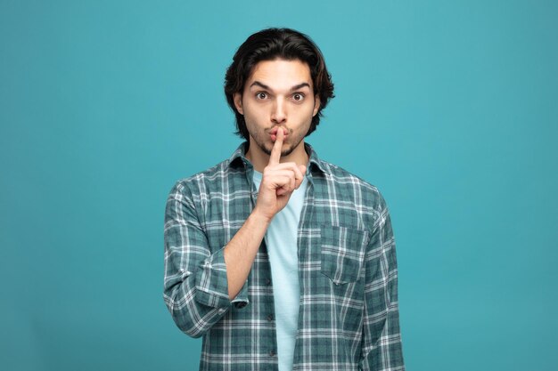 impressed young handsome man looking at camera showing silence gesture isolated on blue background