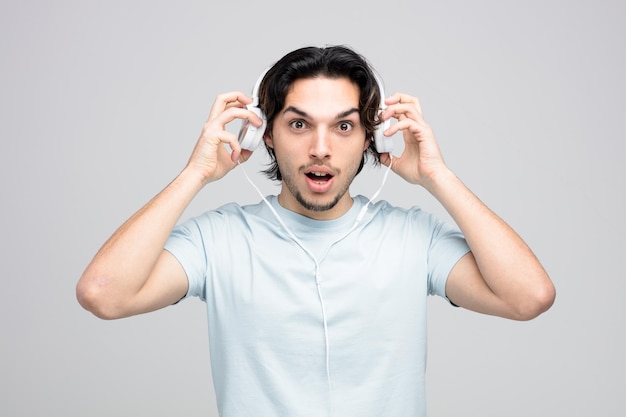 impressed young handsome man looking at camera taking headphones off isolated on white background