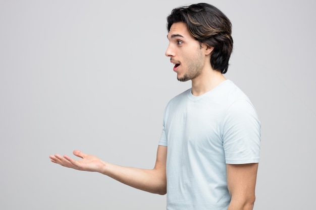 impressed young handsome man standing in profile view looking at side showing empty hand isolated on white background