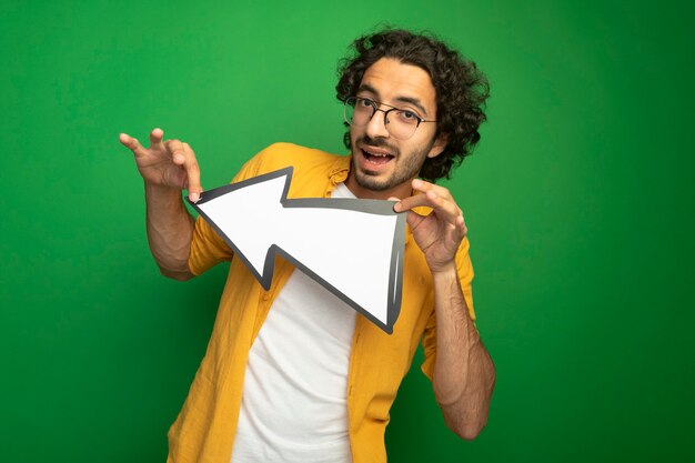 Impressed young handsome man wearing glasses holding arrow mark that is pointing at side looking at front isolated on green wall