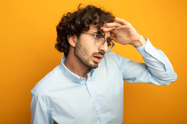 Impressed young handsome man wearing glasses keeping hand on forehead looking at side into distance isolated on orange wall