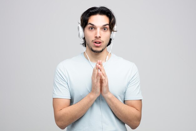 impressed young handsome man wearing headphones keeping hands together looking at camera isolated on white background