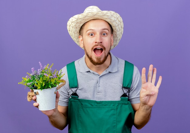 Бесплатное фото Впечатленный молодой красивый славянский садовник в униформе и шляпе, держащий цветочный горшок, показывая пустую руку, изолированную на фиолетовой стене