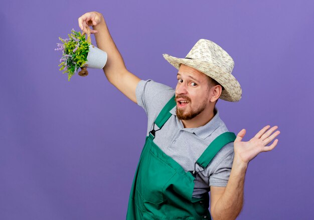 Впечатленный молодой красивый славянский садовник в униформе и шляпе, держащий цветочный горшок, глядя, показывая пустую руку, изолированную на фиолетовой стене