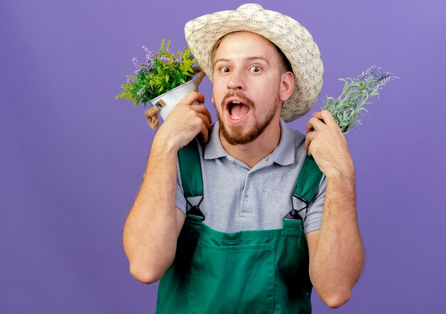 Впечатленный молодой красивый славянский садовник в форме и шляпе, держащий цветочные горшки на плечах, смотрит