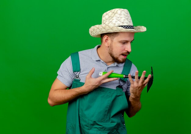 Impressed young handsome slavic gardener in uniform and hat holding and looking at hoe-rake 