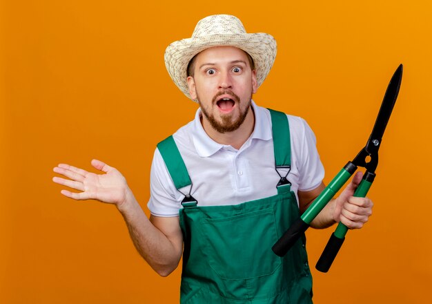 Impressed young handsome slavic gardener in uniform and hat holding pruners showing empty hand isolated on orange wall