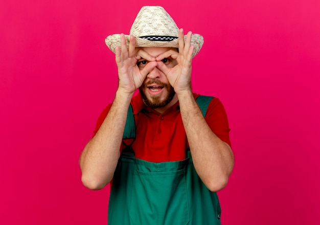 Foto gratuita impressionato giovane giardiniere slavo bello in uniforme e cappello che osserva facendo gesto di sguardo usando le mani come binocolo