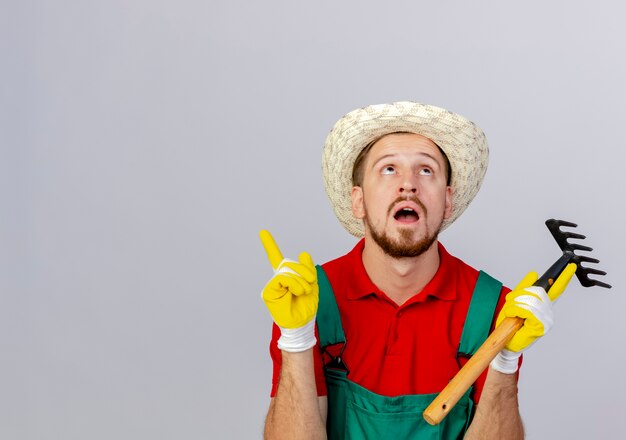 Impressed young handsome slavic gardener in uniform and hat wearing gardener gloves holding rake looking and pointing up isolated