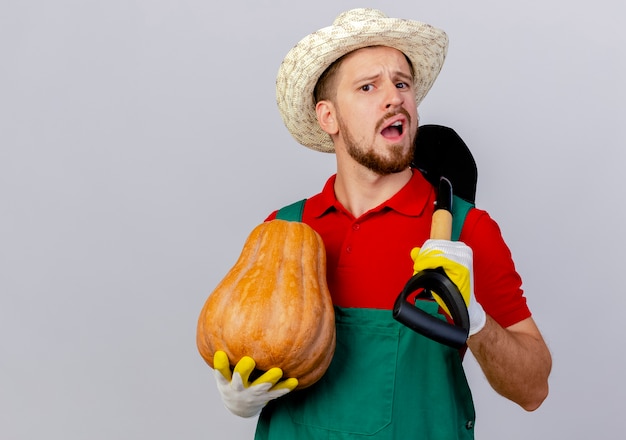 Free photo impressed young handsome slavic gardener in uniform wearing gardening gloves and hat holding butternut pumpkin and spade looking isolated