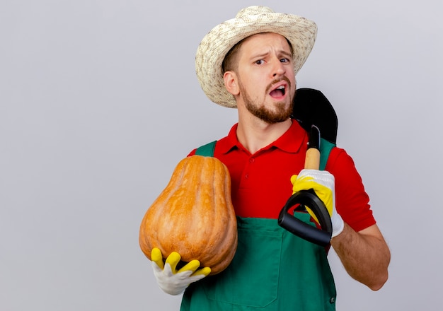 Impressed young handsome slavic gardener in uniform wearing gardening gloves and hat holding butternut pumpkin and spade looking isolated