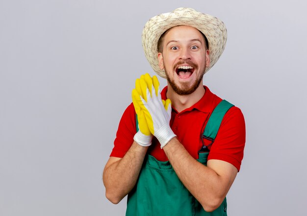 Impressed young handsome slavic gardener in uniform wearing gardening gloves and hat looking keeping hands together isolated