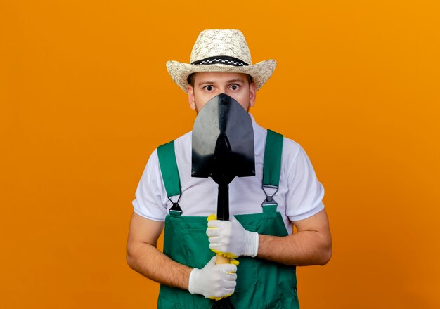 Impressed young handsome slavic gardener in uniform wearing hat and gardening gloves holding spade looking from behind it isolated