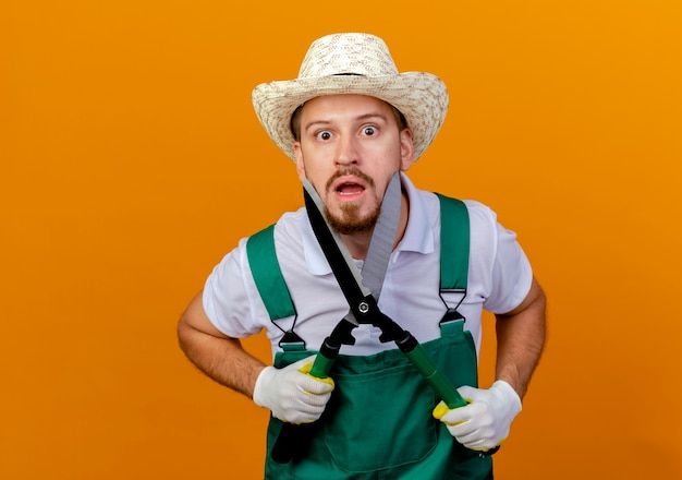 Impressed young handsome slavic gardener in uniform wearing hat and gardening gloves touching face with pruners looking isolated