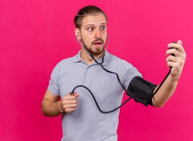 Impressed young handsome slavic ill man wearing stethoscope measuring his pressure with sphygmomanometer looking at it isolated on pink wall with copy space