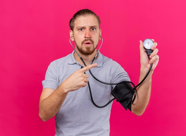 Impressed young handsome slavic ill man wearing stethoscope measuring his pressure with sphygmomanometer pointing at it looking at front isolated on pink wall with copy space